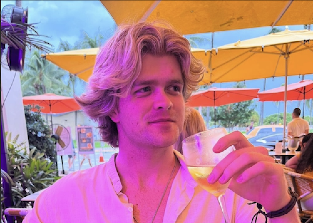 Matt sipping a white wine with table umbrellas behind him. The image hue has been manipulated with some hues of pink.